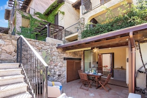 A stone staircase leading to a wooden deck with outdoor furniture, a stone wall, and a building with balconies and greenery.