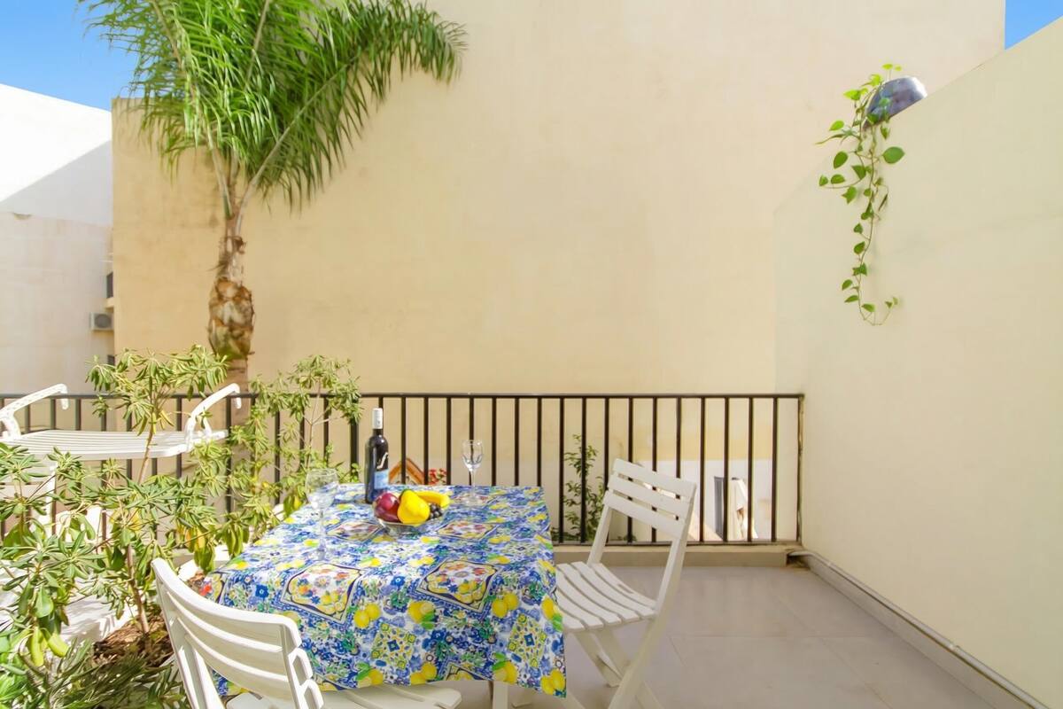Un pequeño espacio de comedor al aire libre con una mesa cubierta con un mantel estampado, rodeada de sillas blancas, una planta en maceta y una barandilla.