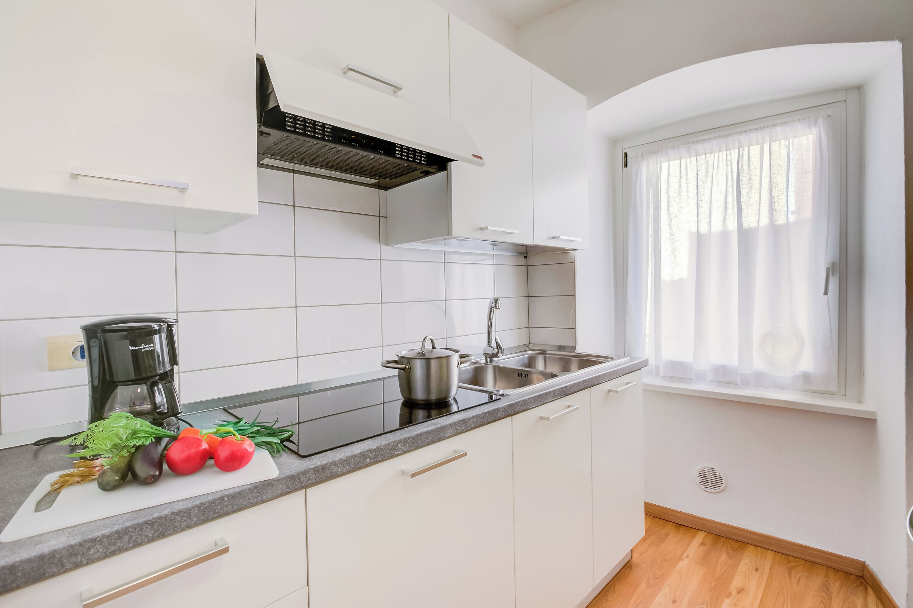 A modern kitchen with white cabinets, a black countertop, a coffee maker, a pot, and fresh vegetables.