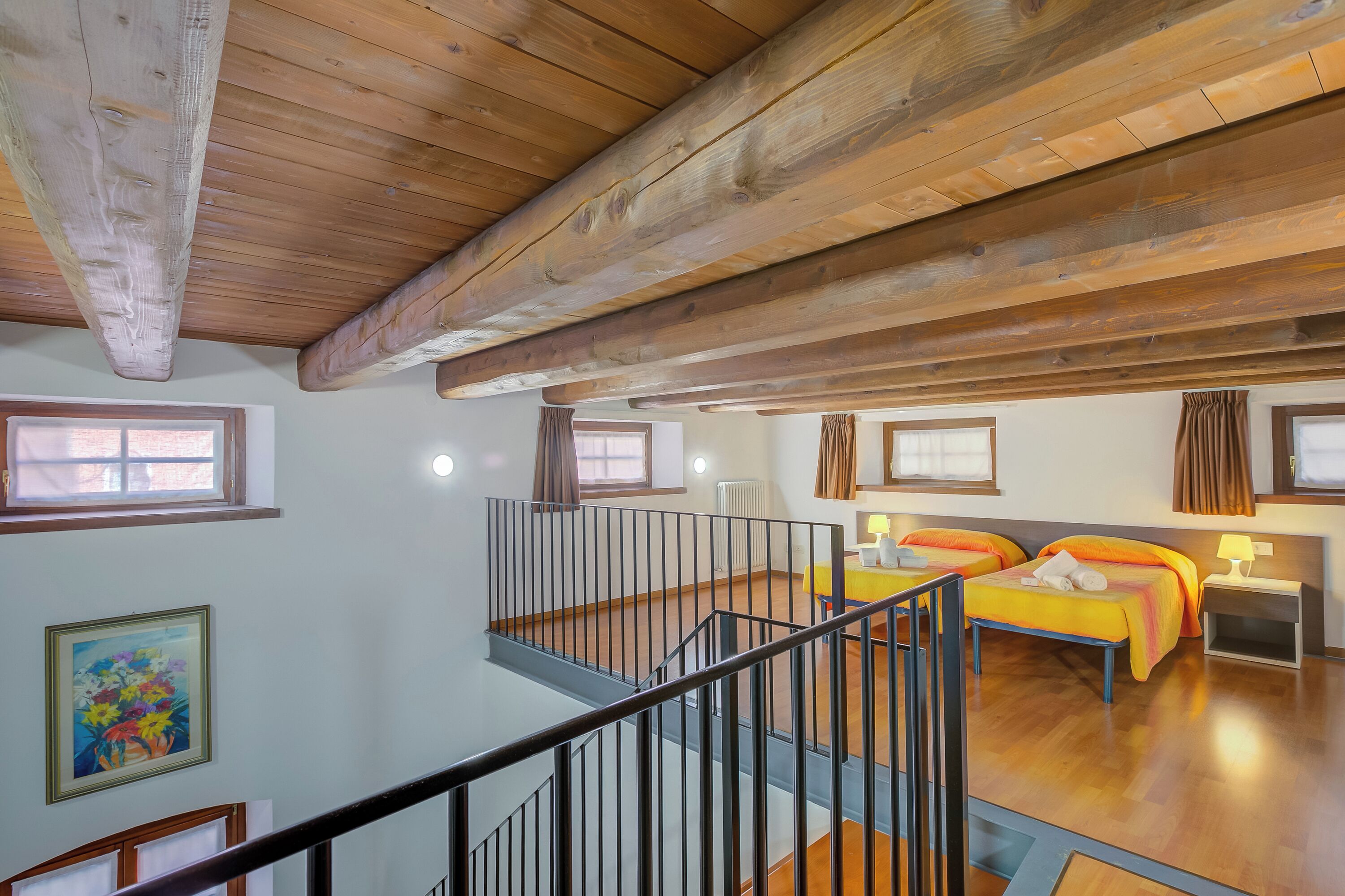 A spacious room with wooden beams on the ceiling, two beds with orange bedding, a small table, and a window with curtains.