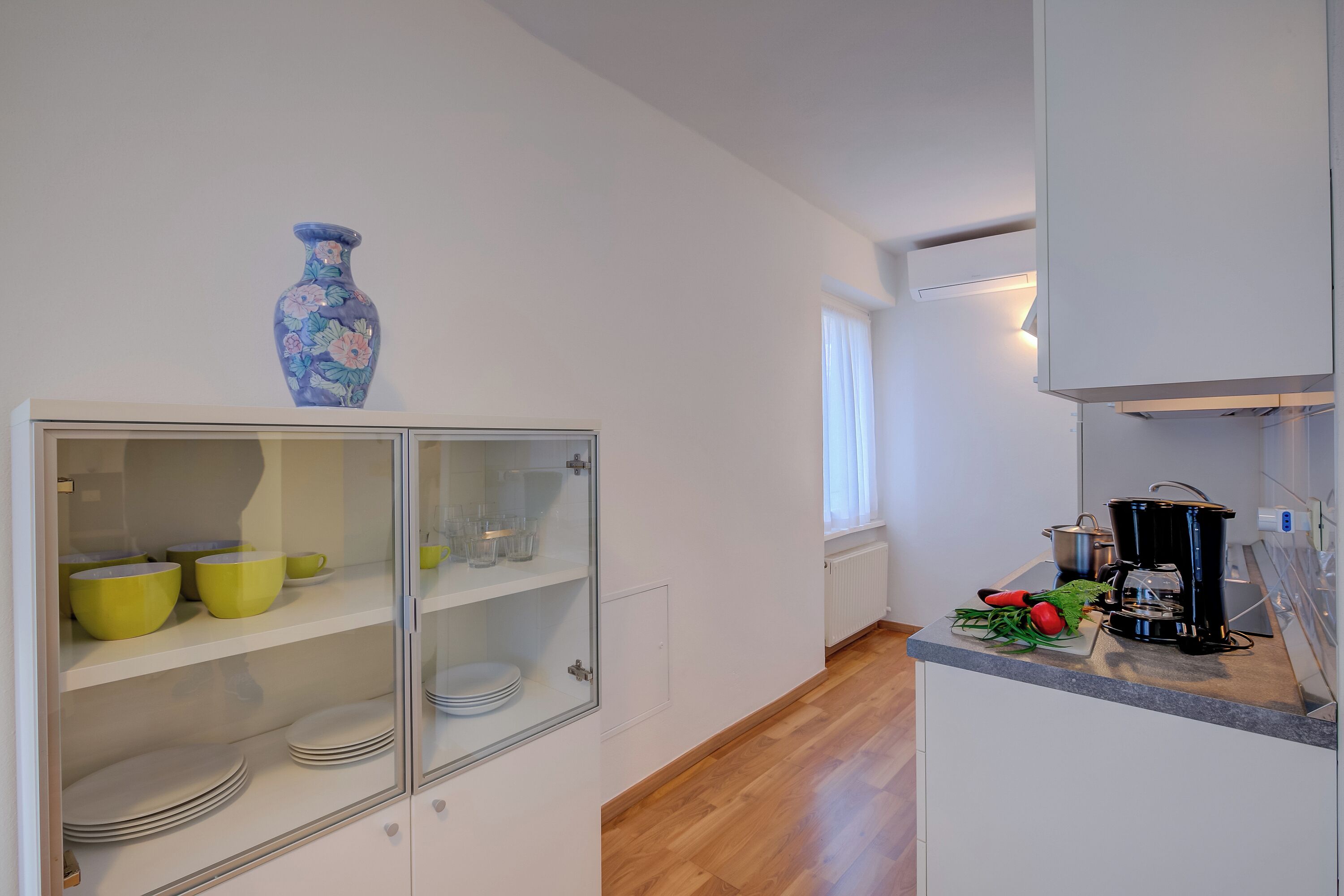 A kitchen with white cabinets, a glass-fronted cabinet with yellow bowls, a blue vase, and a coffee maker on the counter.