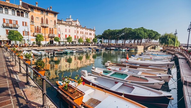 Un port de plaisance où les bateaux sont amarrés le long d’une promenade en bord de mer, entouré de bâtiments historiques et d’arbres.