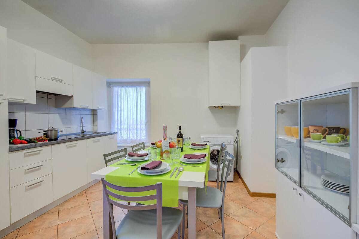 Une cuisine moderne avec des meubles blancs, une table à manger dressée avec des assiettes, des verres et des couverts, et une fenêtre équipée de stores.