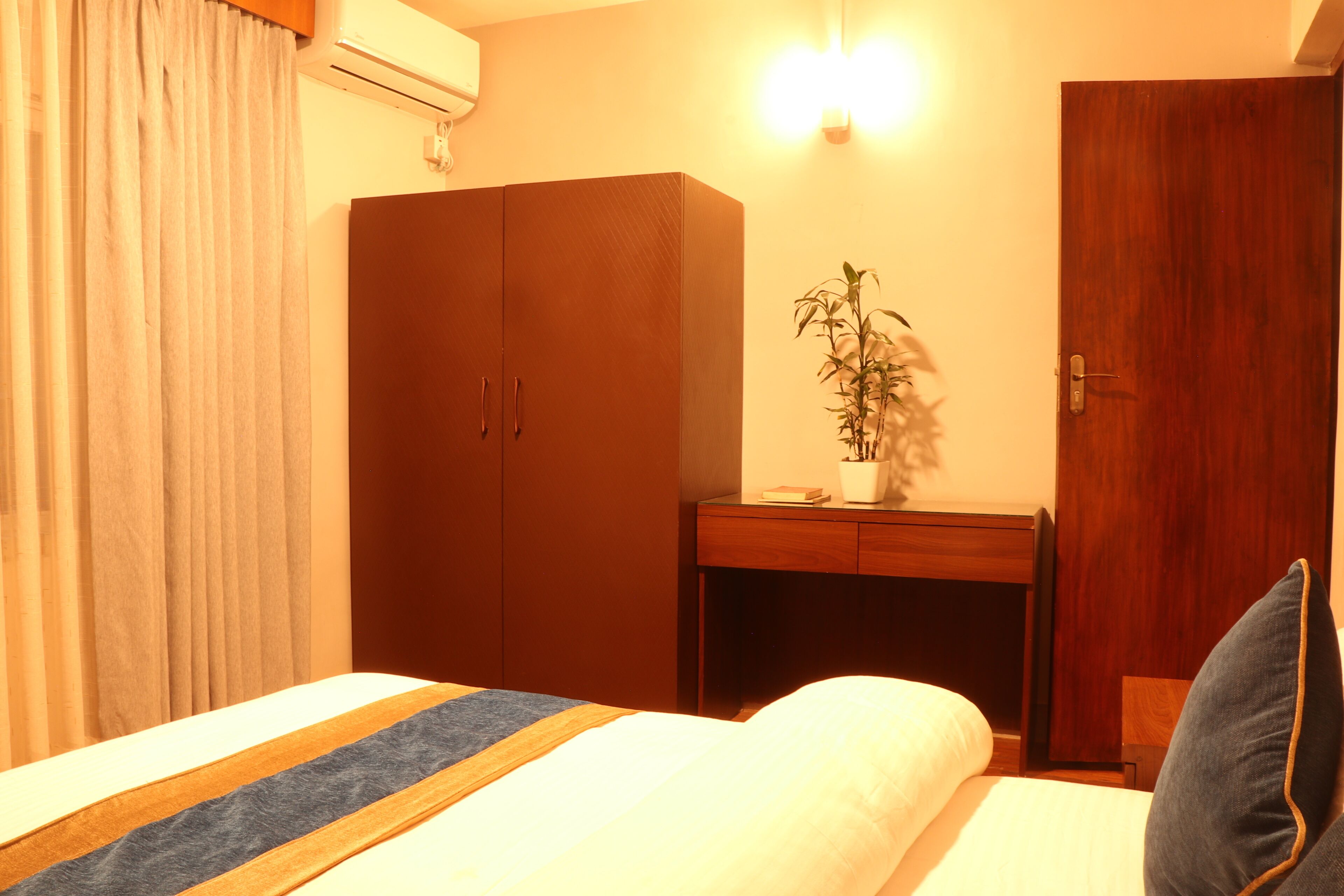 A hotel room with a bed, a wooden wardrobe, a desk with a plant, and a wall-mounted air conditioner.