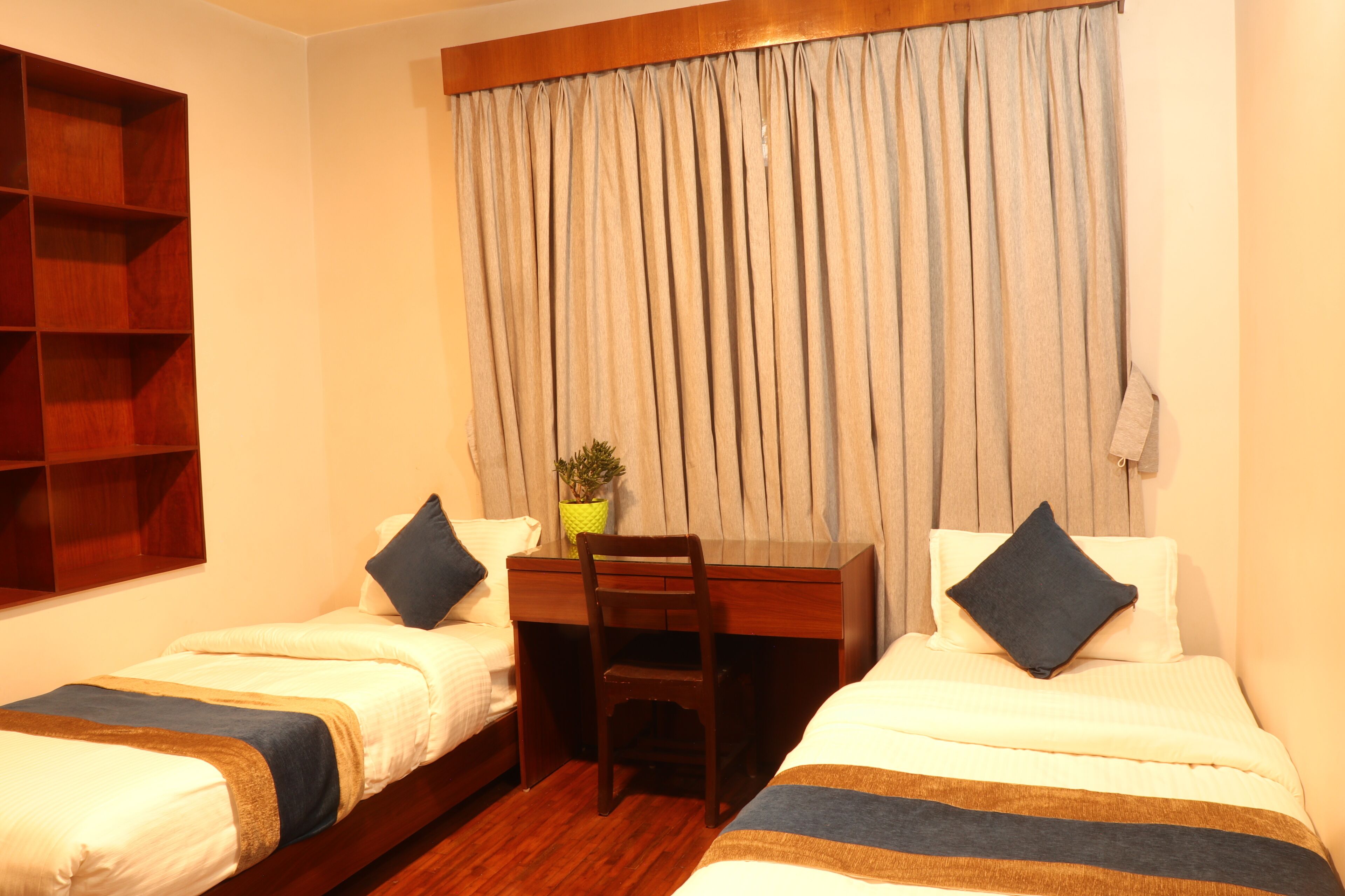 A hotel room with two single beds, a wooden desk, a chair, and a large window with curtains.