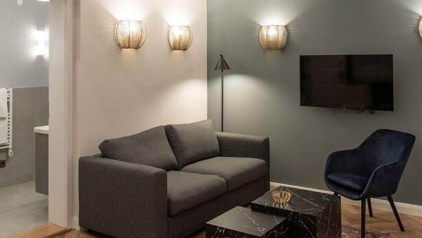 Una sala de estar moderna con un sofá gris, una mesa de centro negra y un sillón azul. Hay un televisor montado en la pared y una escalera que conduce a un nivel superior.