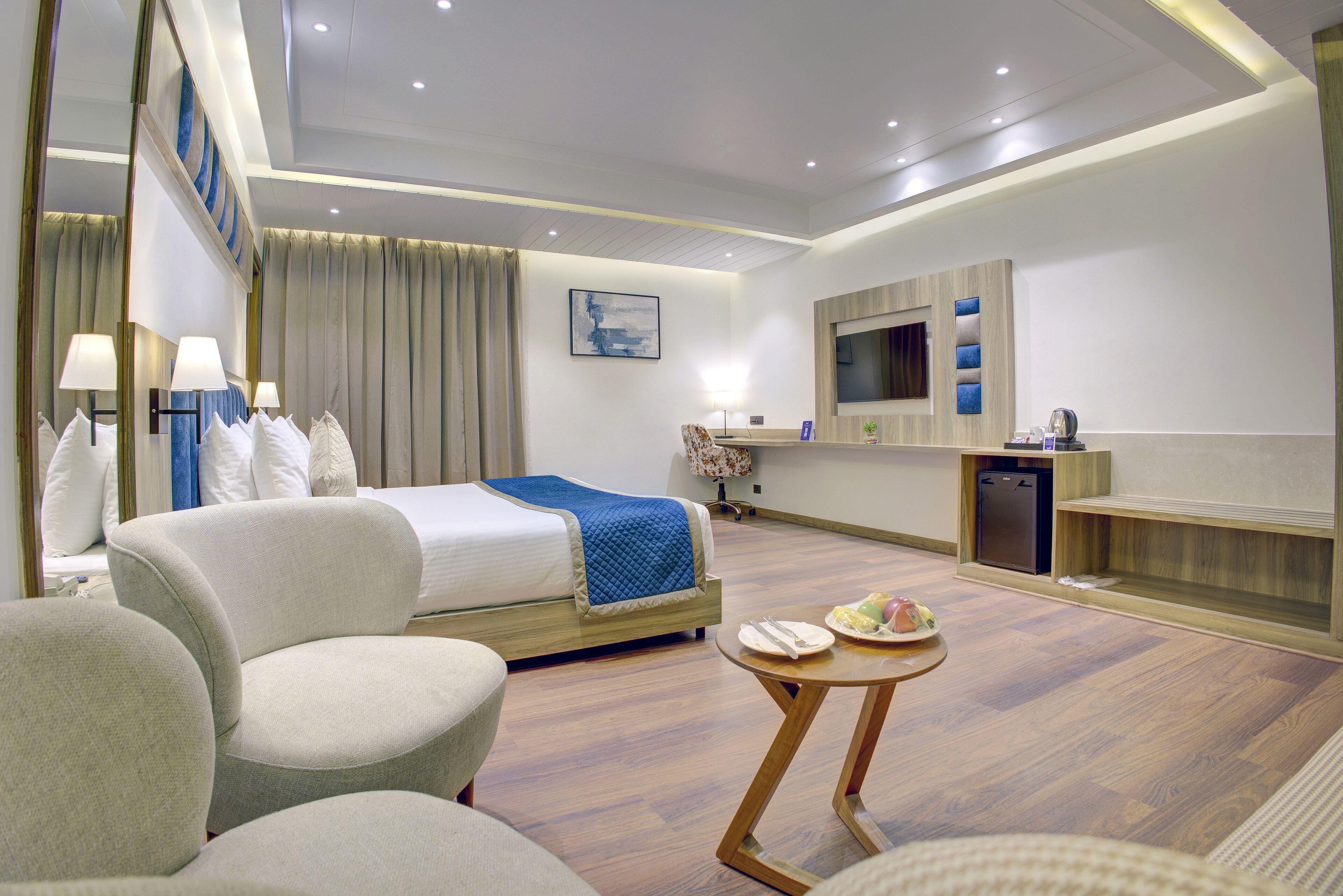 Ein Hotelzimmer mit einem Bett, zwei Sesseln, einem kleinen Tisch mit Essen, einem Fernseher und einem Schreibtisch mit einem Computer.