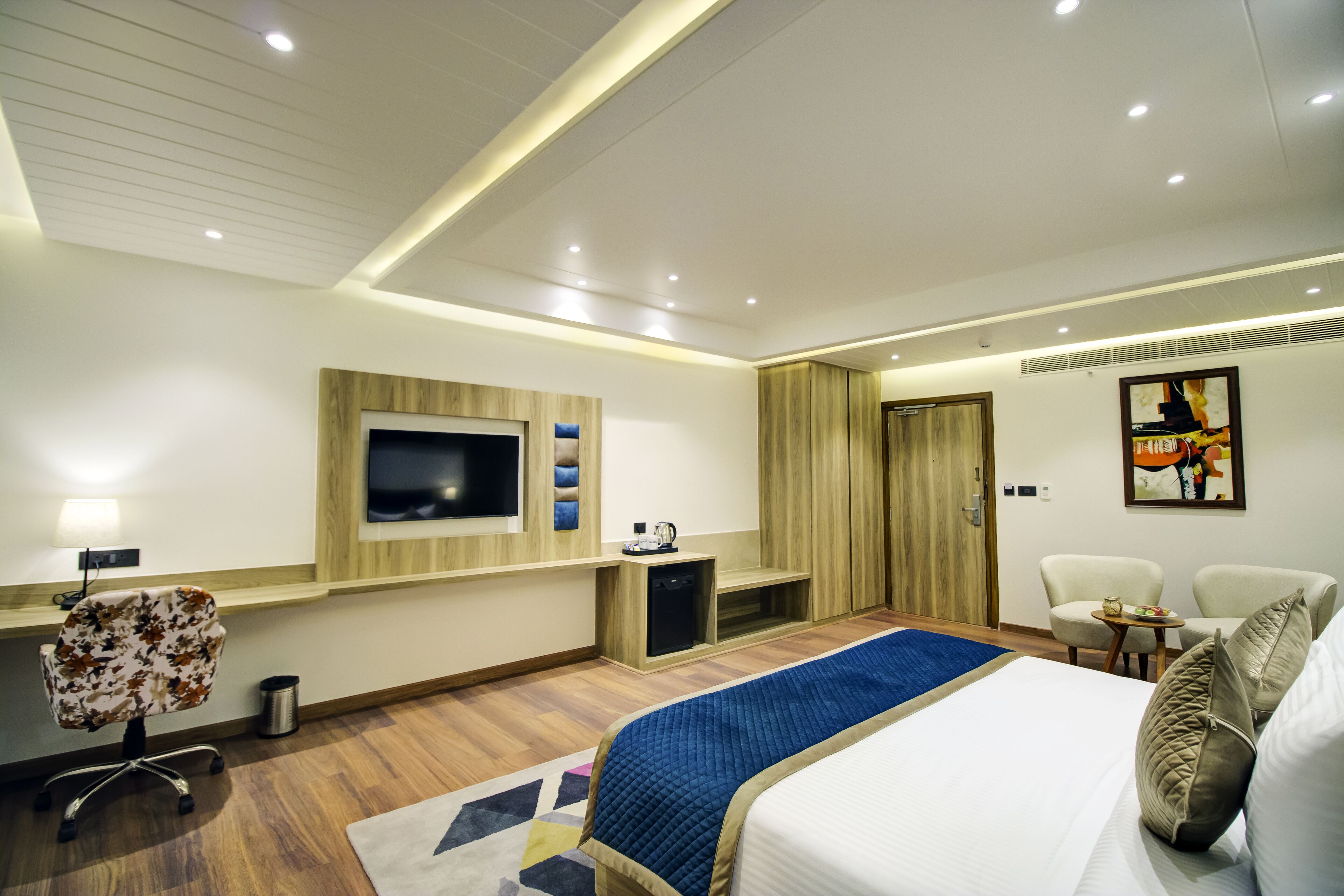 A modern hotel room with a large bed, a TV mounted on the wall, a desk with a chair, and a small table with a lamp.