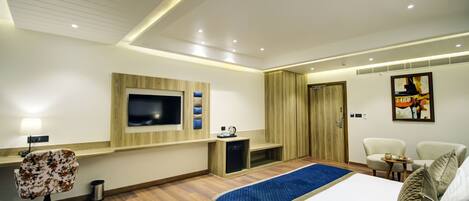 A modern hotel room with a large bed, a TV mounted on the wall, a desk with a chair, and a small table with a lamp.