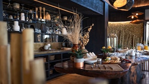 Una zona de bar con estanterías llenas de copas, una barra de madera con un jarrón de plantas secas y una variedad de alimentos y bebidas.