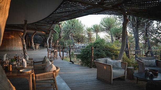 Un espacio de comedor al aire libre con mesas y sillas de madera, rodeado de árboles y una terraza de madera.