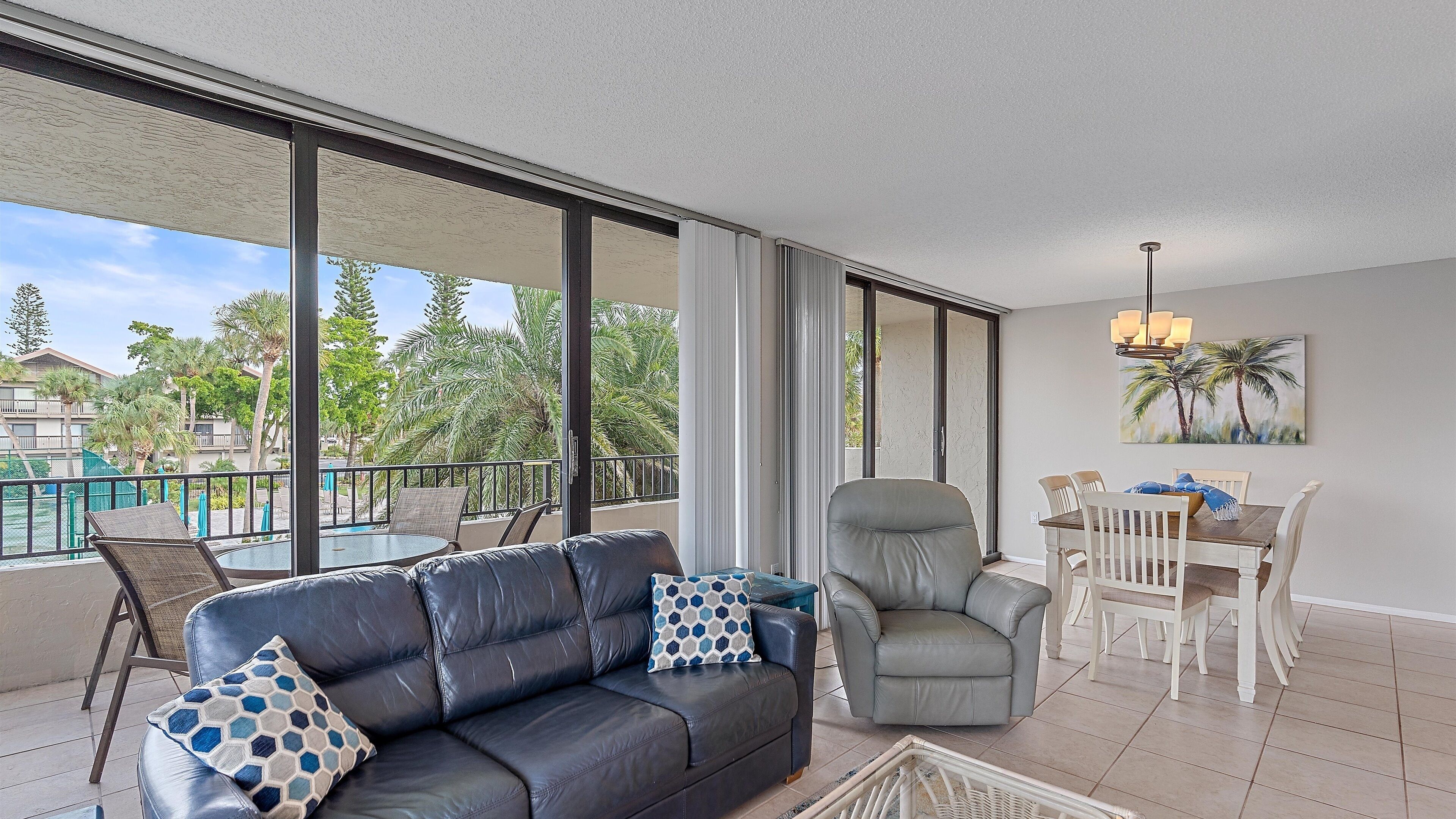 Un vaste espace de vie comprenant un canapé en cuir, un fauteuil gris et une table à manger avec des chaises. De grandes fenêtres offrent une vue sur les palmiers et une piscine.