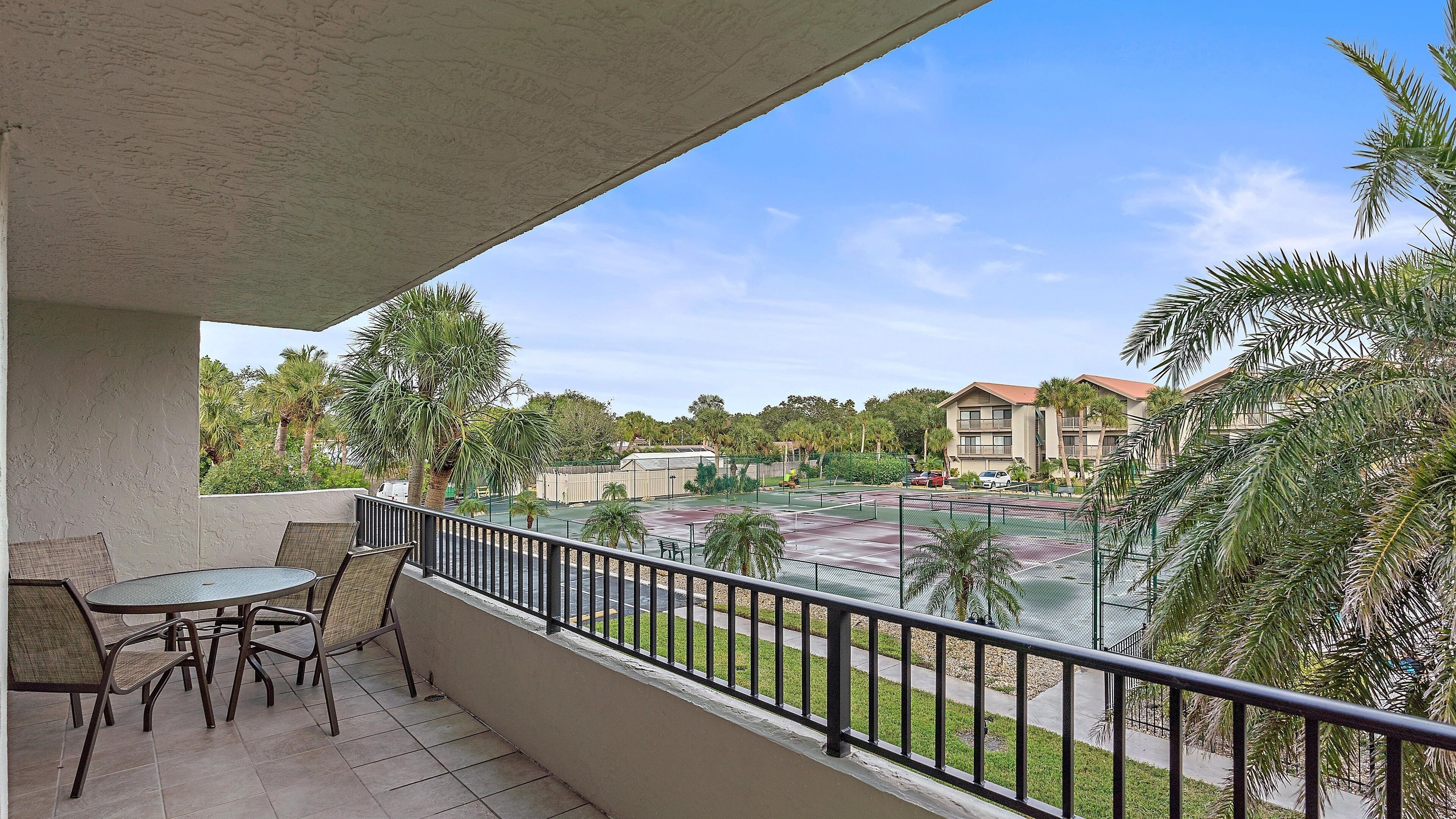 A balcony with a table and chairs, overlooking a tennis court and a building with a red roof.