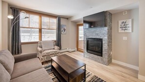 A modern living room with a fireplace, a sofa, a coffee table, and a large window with blinds. - Premium Unit 1307 - 1 Bedroom - Zephyr Mountain 1 Condo (Winter Park)