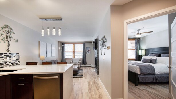 A modern kitchen with a central island, a dining area with a sofa and table, and a bedroom with a bed and nightstand. - Premium Unit 1307 - 1 Bedroom - Zephyr Mountain 1 Condo (Winter Park)