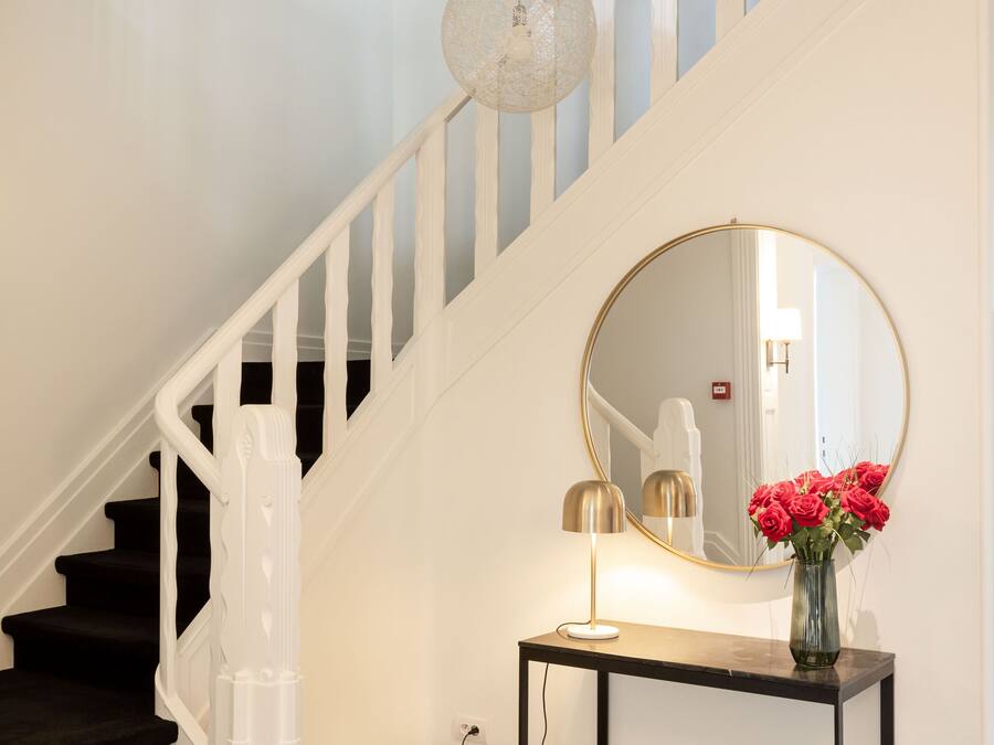 A staircase with black and white checkered flooring, a round mirror on a console table, and a vase with red flowers.