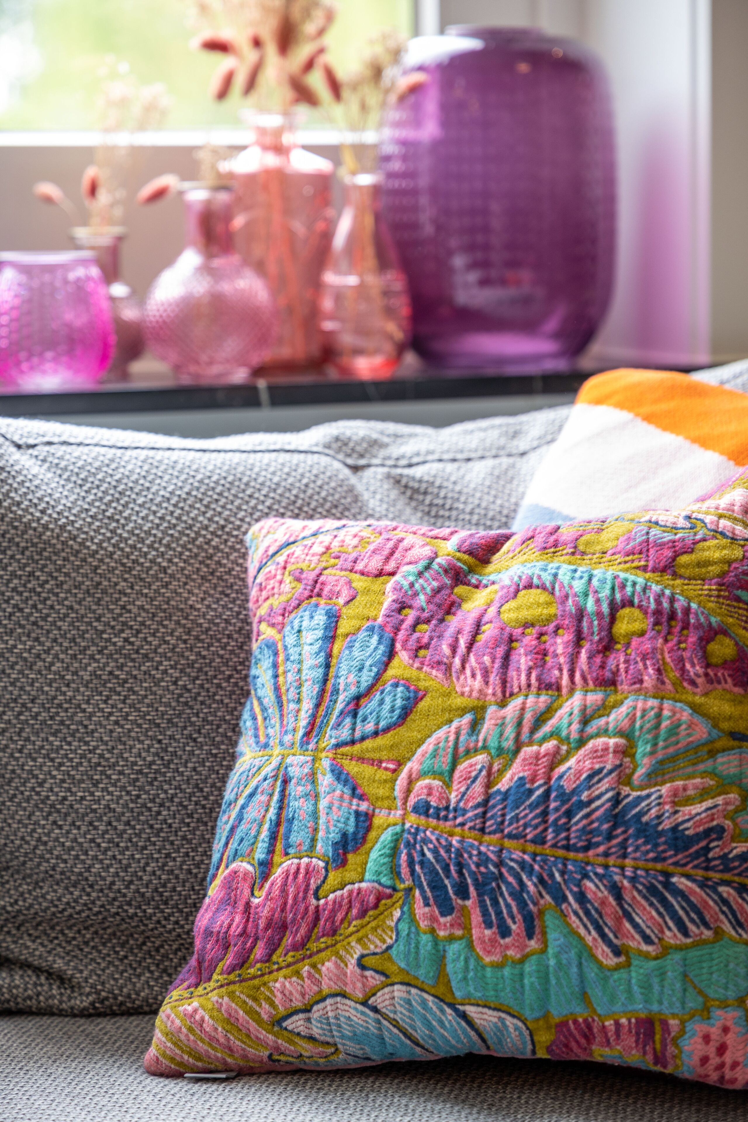 A colorful pillow on a gray couch with a window view of pink and purple vases.