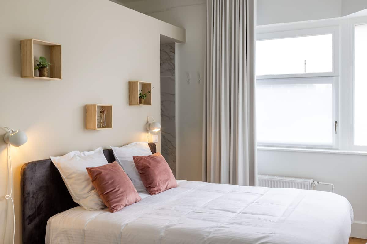 A bedroom with a bed, bedside lamps, a window, and a wall-mounted shelf with a plant.