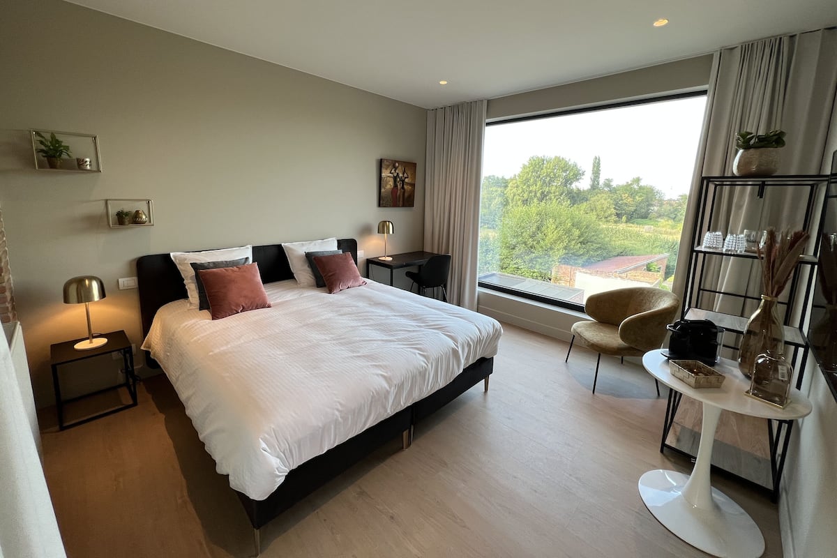 A modern bedroom with a large bed, bedside tables, a desk, a chair, and a view of greenery outside.