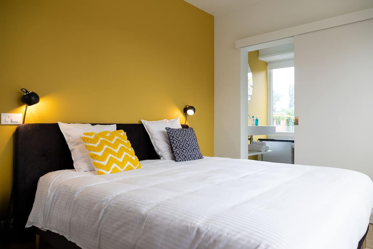 A neatly made bed with white bedding and two patterned pillows, a black lamp, and a mirror above the bed.