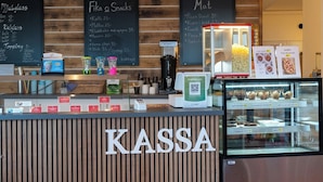 Un mostrador de KASSA con un panel de menú que enumera artículos como muffins y café, una máquina de café y una vitrina con pasteles.