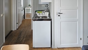 Cocina compacta con refrigerador, estufa y gabinetes blancos. Suelo de madera y una silla en primer plano.