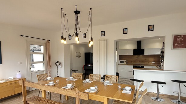 Un comedor con mesa de madera dispuesta para cuatro personas, rodeado de sillas de mimbre. Un candelabro cuelga encima, y se ve una cocina al fondo.