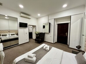 A hotel room with a bed, a seating area with a chair, a TV, a mirror, and a wardrobe.