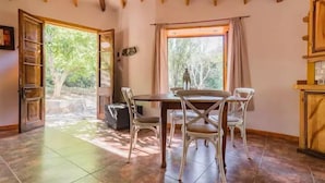 Zona de menjador amb taula i cadires de fusta, una finestra amb cortines i vistes a vegetació exterior.