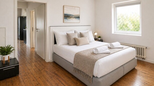 A bedroom with a bed, a window, a radiator, and a door leading to a kitchen.