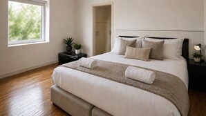 A bedroom with a large bed, a nightstand with a lamp, a potted plant, and a view of greenery outside.