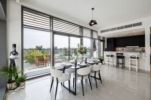 A modern kitchen with a large window, a dining area with white chairs, and a view of the city. - Corner Unit Mulberry Dubai Hills Estate (Dubai)