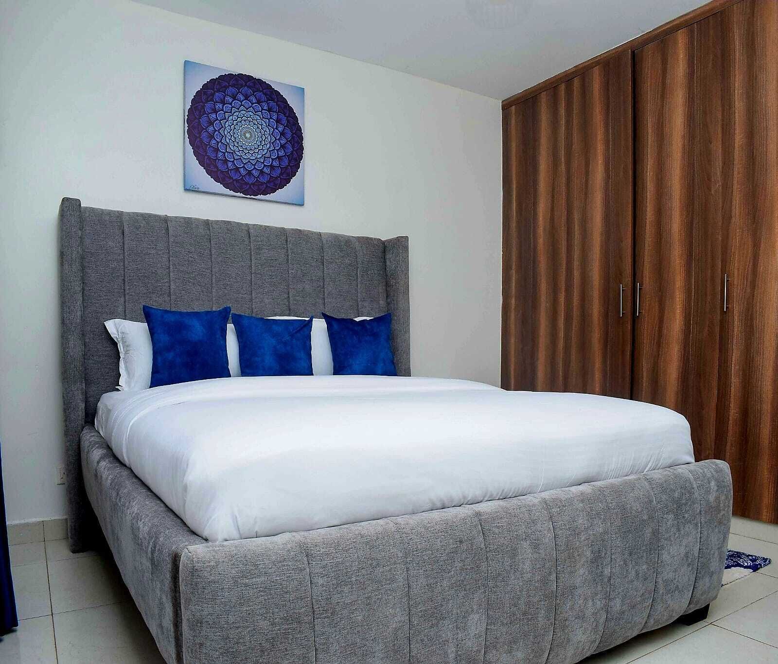 A bedroom with a grey upholstered bed, white bedding, and blue pillows. There is a wooden wardrobe with two doors and a circular artwork on the wall above the bed.