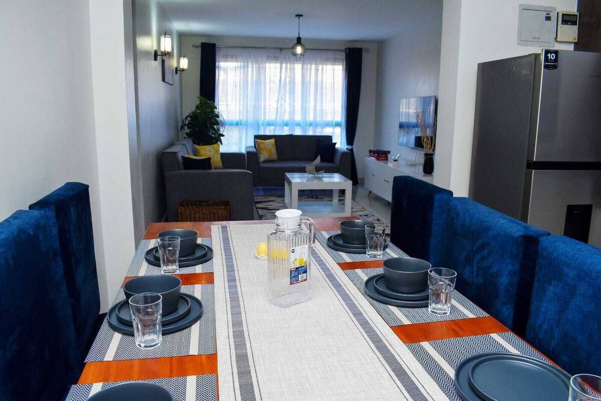 Un comedor con una mesa dispuesta para cuatro personas, un sofá al fondo y una zona de cocina con un refrigerador.