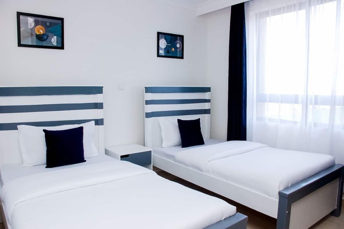 Habitación de hotel con dos camas, cada una con cabecero a rayas, ropa de cama blanca y almohada oscura. Hay dos cuadros en la pared, una ventana con cortinas translúcidas y una mesita de noche entre las camas.