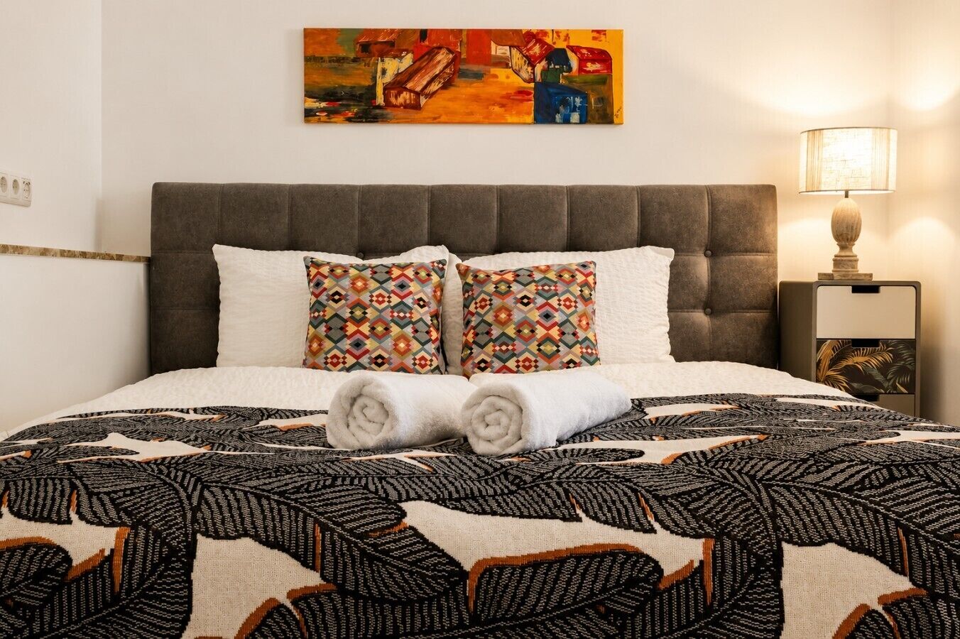 A neatly made bed with patterned pillows, a headboard, a wall painting, and a bedside lamp.