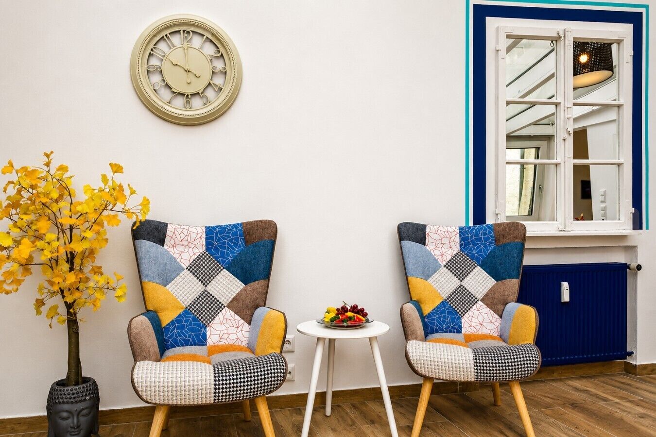 Two armchairs with a patchwork design, a wall clock, a small table with fruit, and a window with a blue frame.