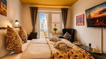 Ein Hotelzimmer mit einem Bett, einem Sessel, einem Fernseher, einem Fenster mit Vorhängen und einem kleinen Tisch mit Pflanze und Obst.