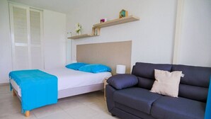 A bedroom with a bed, a blue blanket, a dark sofa, a bedside table with a lamp, and a shelf with decorative items.