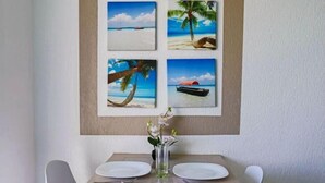 A dining area with a table set for two, featuring a vase with flowers and a framed beach scene above.