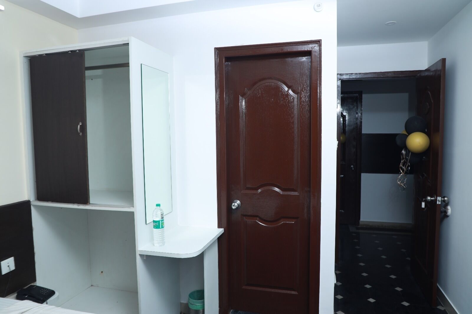 Habitación de hotel con puerta de madera, pared blanca, espejo y estante con una botella de agua.