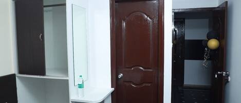 Habitación de hotel con puerta de madera, pared blanca, espejo y estante con una botella de agua.