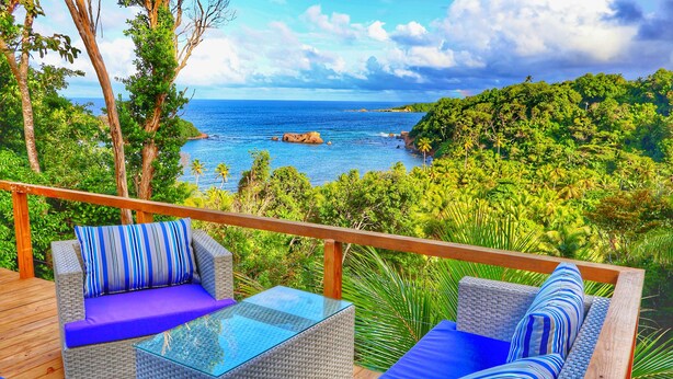 A deck with wicker furniture and blue cushions, overlooking a tropical landscape with a view of the ocean. - Blue Whale Villa - Penthouse (Calibishie)