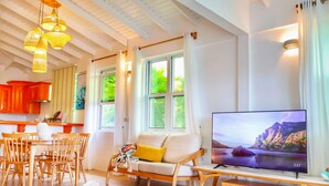 A modern living room with a sofa, dining table, and chairs, a flat-screen TV, and a kitchen area with orange cabinets. - Blue Whale Villa - Penthouse (Calibishie)