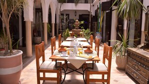 Un espace de restauration en plein air avec une table dressée pour deux personnes, entouré de plantes en pot et d’une fontaine.