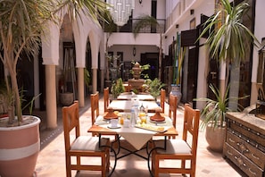 An outdoor dining area with a table set for two, surrounded by potted plants and a fountain. - Riad Kalé Polis (Marrakech)