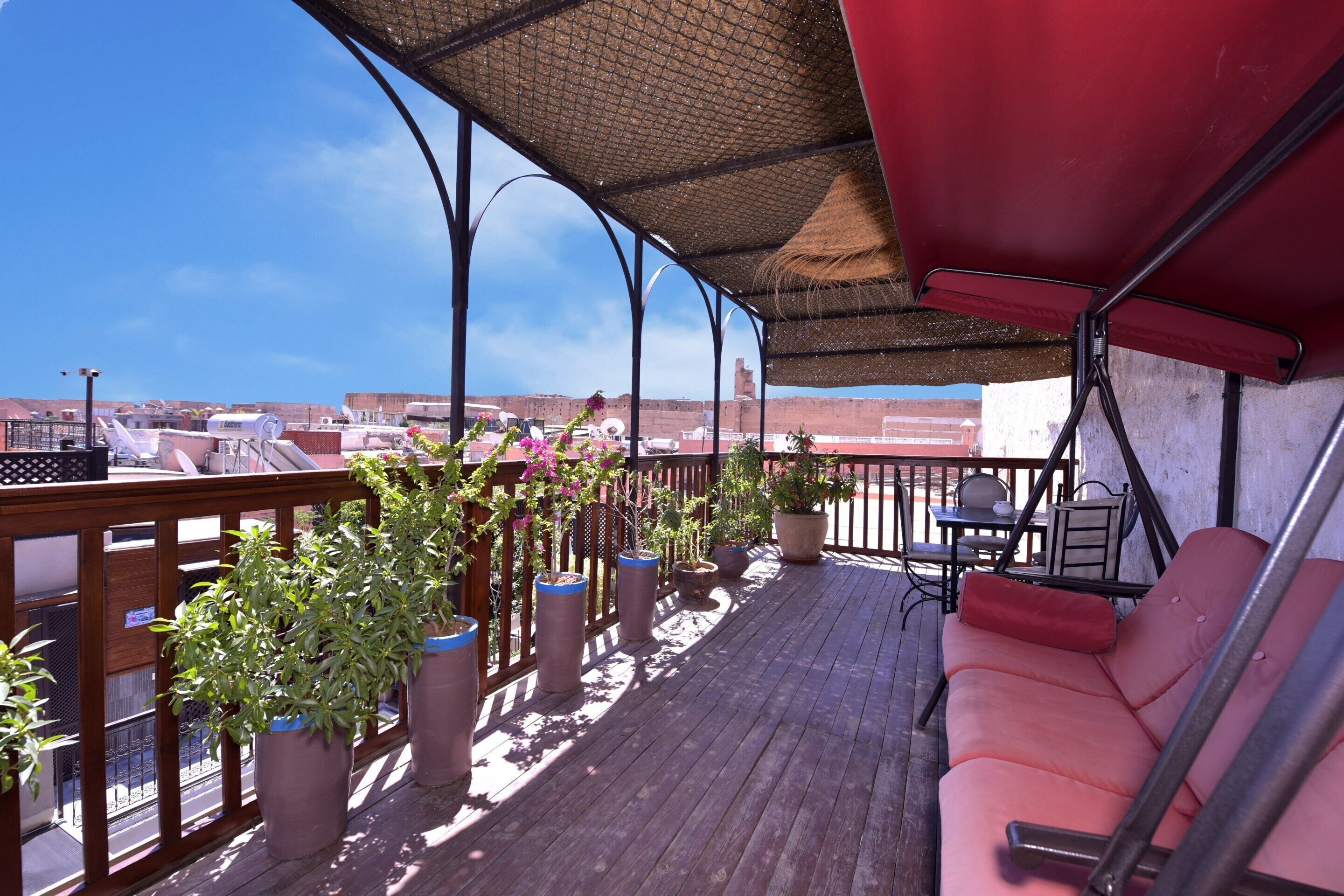 Um deck coberto ao ar livre com piso de madeira, plantas em vasos, um toldo vermelho e uma área de estar com mesa e cadeiras.