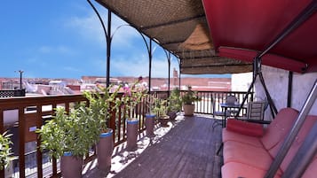 Um deck coberto ao ar livre com piso de madeira, plantas em vasos, um toldo vermelho e uma área de estar com mesa e cadeiras.