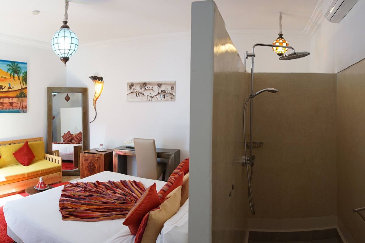 A hotel room with a bed, a desk, a chair, a shower, and artwork on the walls.