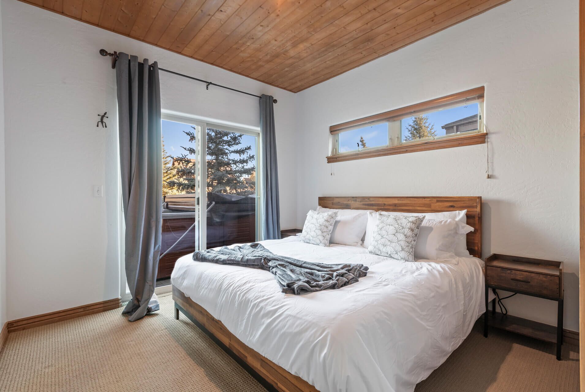 Unwind in your serene bedroom with warm wood accents, plush bedding, and natural light streaming through windows that frame views of evergreen trees.